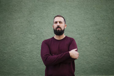 Bearded man with arms crossed standing against green wall - MIMFF00384
