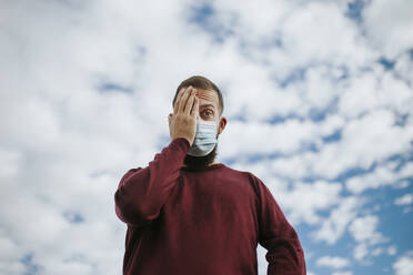 Mid adult man wearing mask hiding face with hand while standing against cloudy sky - MIMFF00388