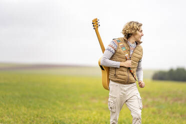 Junger Mann geht mit Akustikgitarre auf dem Rücken über eine Wiese - GGGF00638