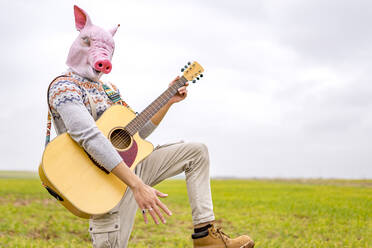 Porträt eines jungen Mannes mit Schweinemaske, der auf einer Wiese Gitarre spielt - GGGF00656