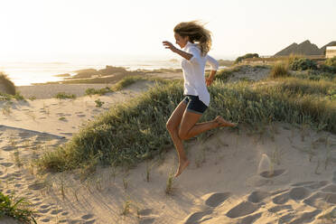 Carefree woman jumping on sand at beach during sunset - SBOF02322