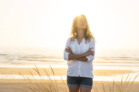 Mid adult woman standing wit arms crossed at beach during sunset stock photo