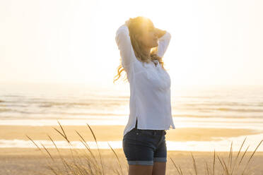 Carefree woman with hands behind head standing at beach - SBOF02329