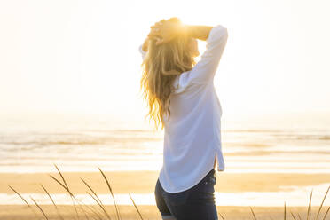 Woman with hands behind head standing at beach during sunset - SBOF02330