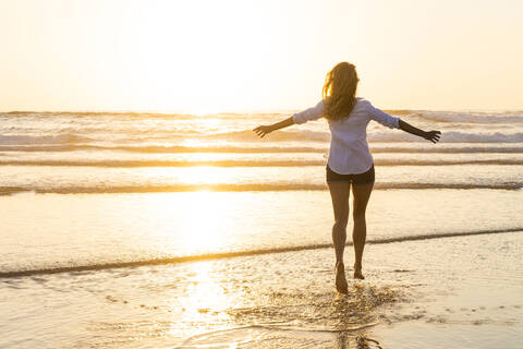 Carefree woman with arms outstretched running toward sea during sunset stock photo