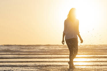 Silhouette einer Frau, die bei Sonnenuntergang am Strand steht und im Wasser plantscht - SBOF02334