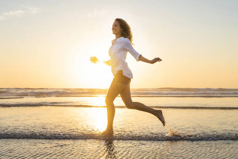 Happy woman looking away while running against sea during sunset - SBOF02335