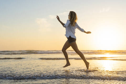 Carefree woman running with arms outstretched against sea during sunset stock photo