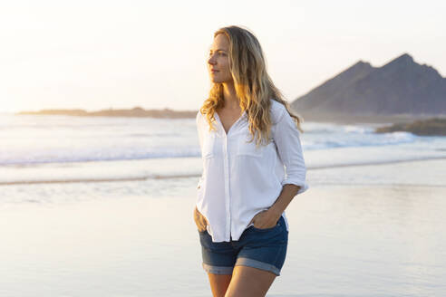 Beautiful woman with hands in pockets looking away while walking at beach - SBOF02340