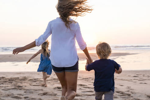 Mutter hält die Hand ihres Sohnes, während sie mit ihrer Tochter im Hintergrund am Strand läuft - SBOF02353