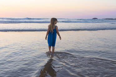 Mädchen in blauem Kleid mit Blick auf die Aussicht beim Spaziergang am Strand bei Sonnenuntergang - SBOF02361