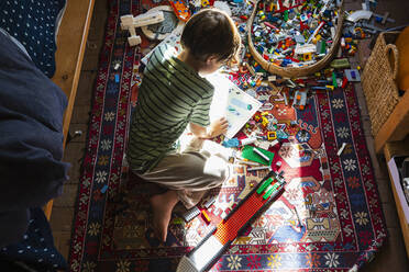 Junge sitzt zwischen Spielzeug auf dem Fußboden seines Schlafzimmers in einem Fleckchen Sonnenlicht - MINF15465