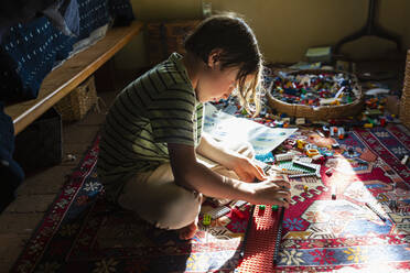 Junge sitzt zwischen Spielzeug auf dem Fußboden seines Schlafzimmers in einem Fleckchen Sonnenlicht - MINF15467