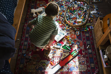 Junge sitzt zwischen Spielzeug auf dem Fußboden seines Schlafzimmers in einem Fleckchen Sonnenlicht - MINF15468