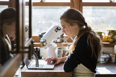 Teenage girl in a kitchen following a baking recipe on a laptop. - MINF15473