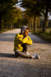 Sportswoman wearing hooded shirt looking away while sitting on road - MAUF03720