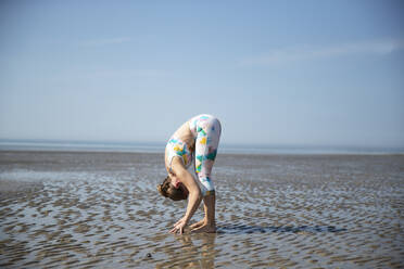 Ältere Frau beugt sich beim Yoga am Strand gegen den Himmel - PMF01693