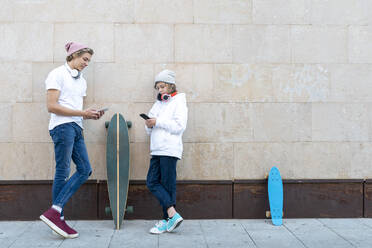 Junger Mann und Junge mit Strickmütze, der ein Mobiltelefon benutzt, während er mit dem Skateboard an der Wand steht - GGGF00660