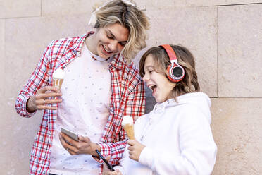 Smiling man with ice cream using mobile phone while standing by boy against wall - GGGF00666