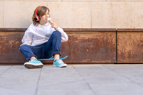 Junge mit Kopfhörern, der auf dem Gehweg sitzend ein Eis isst - GGGF00669