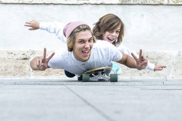 Lächelnde Freunde gestikulieren das Friedenszeichen, während sie auf einem Skateboard auf dem Gehweg liegen - GGGF00674