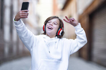 Smiling boy wearing headphones showing peace gesture while taking selfie through mobile phone standing outdoors - GGGF00683