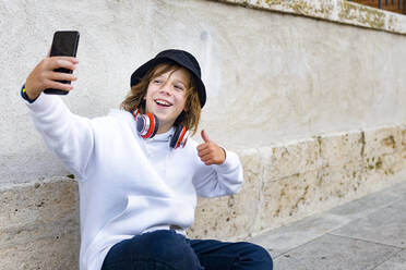 Boy wearing hat and headphones using mobile phone while sitting against wall - GGGF00684