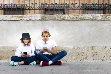 Freunde mit Strickmütze benutzen Mobiltelefone, während sie auf dem Gehweg sitzen - GGGF00686