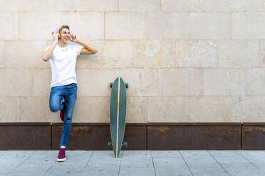 Smiling man with skateboard adjusting headphones while leaning on wall - GGGF00687