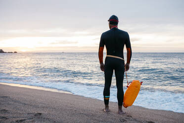 Männlicher Schwimmer, der allein am sandigen Küstenstrand steht, mit Schwimmflügel in der Hand - JRFF04961