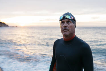 Porträt eines männlichen Schwimmers, der bei Sonnenuntergang allein am Küstenstrand steht - JRFF04964