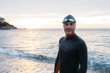 Porträt eines männlichen Schwimmers, der bei Sonnenuntergang allein am Küstenstrand steht - JRFF04967