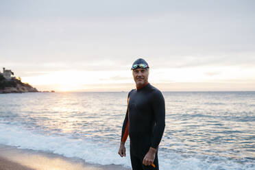 Porträt eines männlichen Schwimmers, der bei Sonnenuntergang allein am Küstenstrand steht - JRFF04968