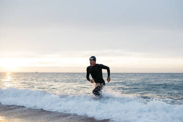 Männlicher Schwimmer verlässt das Wasser nach dem Schwimmen im Meer bei Sonnenuntergang - JRFF04974