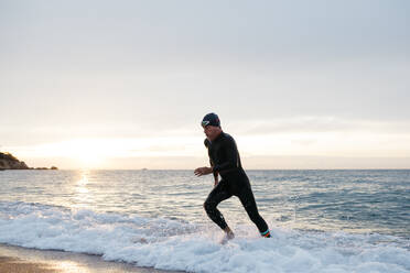 Männlicher Schwimmer verlässt das Wasser nach dem Schwimmen im Meer bei Sonnenuntergang - JRFF04975