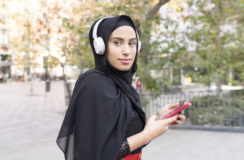 Porträt einer jungen schönen Frau mit Hidschab und Kopfhörern, die ein Smartphone benutzt, lizenzfreies Stockfoto