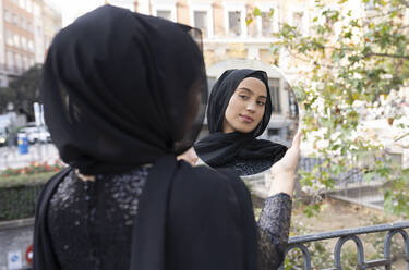Junge schöne Frau mit schwarzem Hidschab steht im Freien und schaut in einen runden Spiegel - JCCMF00473