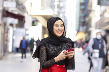 Porträt der jungen schönen Frau mit schwarzem Hijab lächelnd auf dem Bürgersteig mit Smartphone in den Händen - JCCMF00494