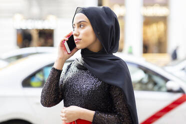 Porträt einer jungen schönen Frau mit schwarzem Hidschab, die mitten auf der Straße mit einem Smartphone telefoniert - JCCMF00501