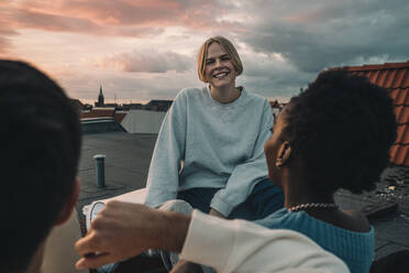 Porträt einer lächelnden Frau, die mit Freunden auf der Terrasse eines Gebäudes bei Sonnenuntergang sitzt - MASF21107