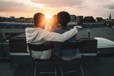 Rückansicht eines Mannes und einer Frau, die bei Sonnenuntergang auf dem Dach eines Gebäudes sitzen - MASF21109