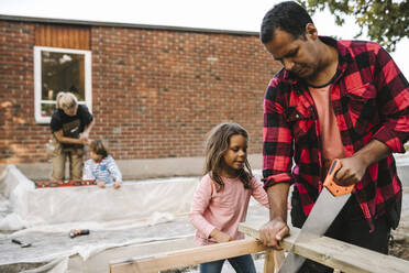 Vater und Tochter arbeiten mit einer Handsäge, während die Mutter mit ihrem Sohn an der Betonwand eines Hauses steht - MASF21120