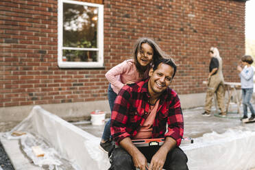 Porträt eines lächelnden, zuversichtlichen Mannes mit Tochter, der an einer Betonwand vor einem Haus sitzt - MASF21121