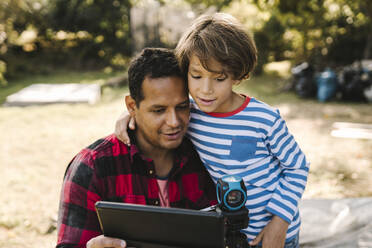 Confident father using digital tablet by son in yard - MASF21152