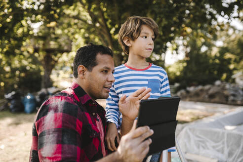 Father with digital tablet gesturing by son in yard - MASF21154