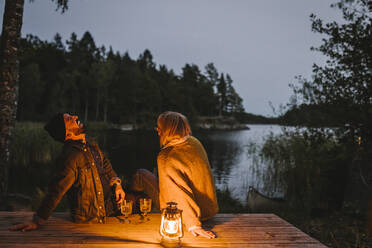 Fröhlicher Mann mit Partnerin sitzt bei Sonnenuntergang auf dem Pier am See - MASF21170