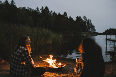 Rückansicht von Freundinnen, die sich bei Sonnenuntergang an einer Feuerstelle am See unterhalten - MASF21171