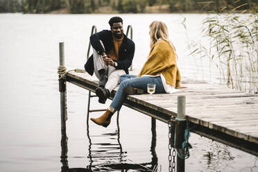 Smiling male and female partners talking while sitting on pier over lake during social gathering - MASF21176