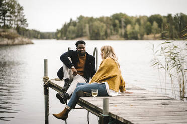 Smiling male and female partners sitting on pier over lake during social gathering - MASF21178