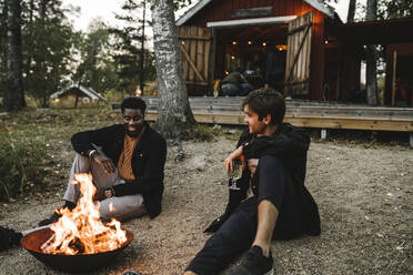 Male friends with drinking glass talking while sitting by fire pit during weekend - MASF21180